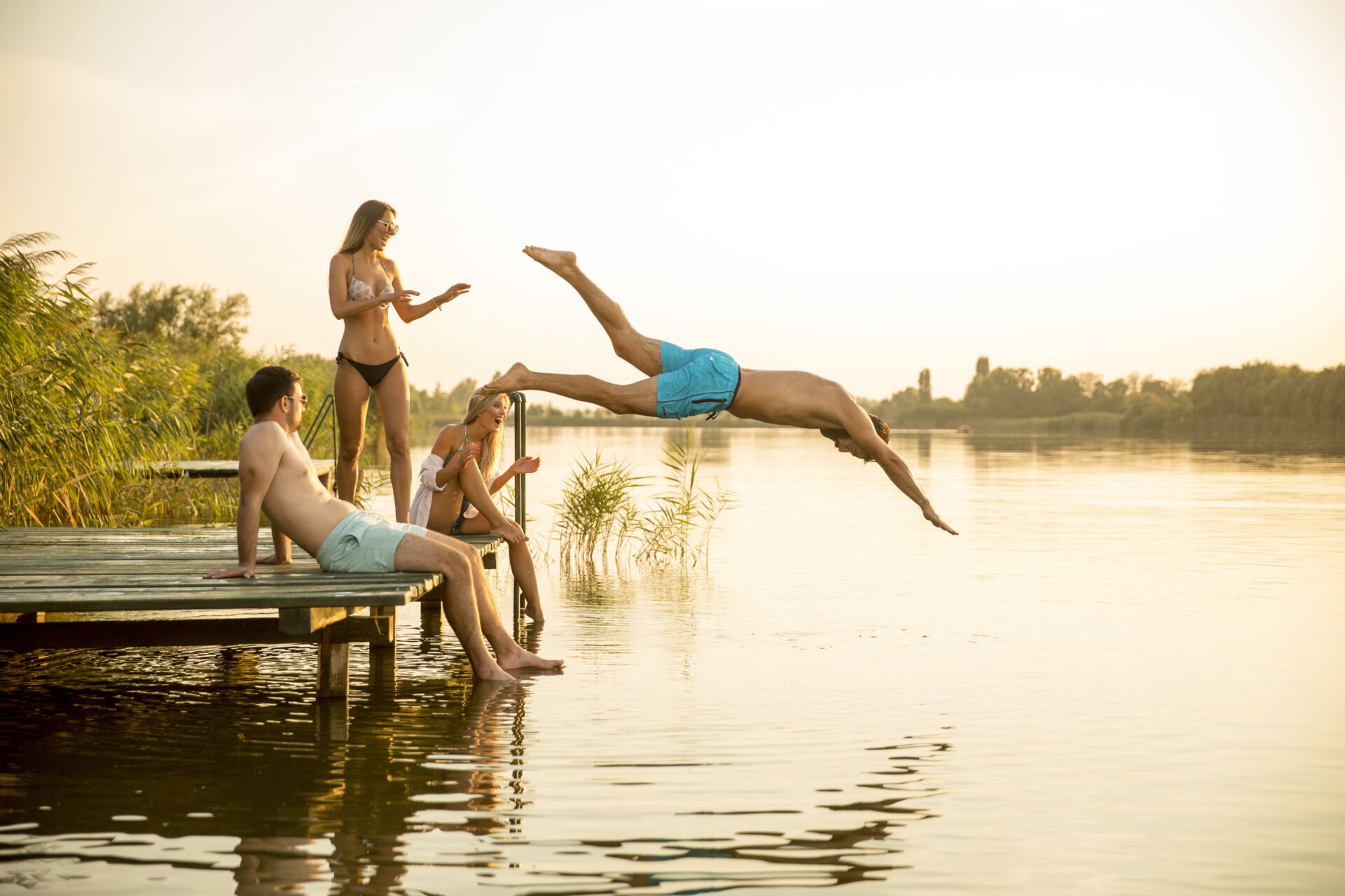 4 Personen die auf einem Steg sitzen, lachen und einer springt in die Masuren