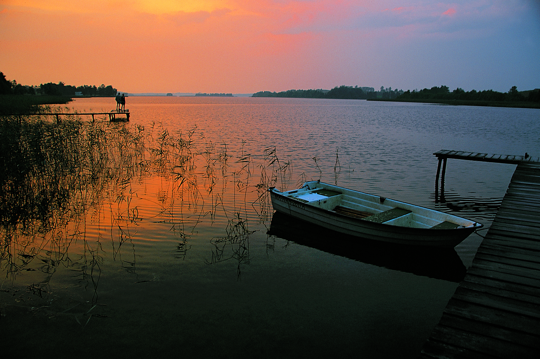Ein See in Masuren bei Sonnenuntergang, mit einem kleinen Boot und einem Steg im Vordergrund.